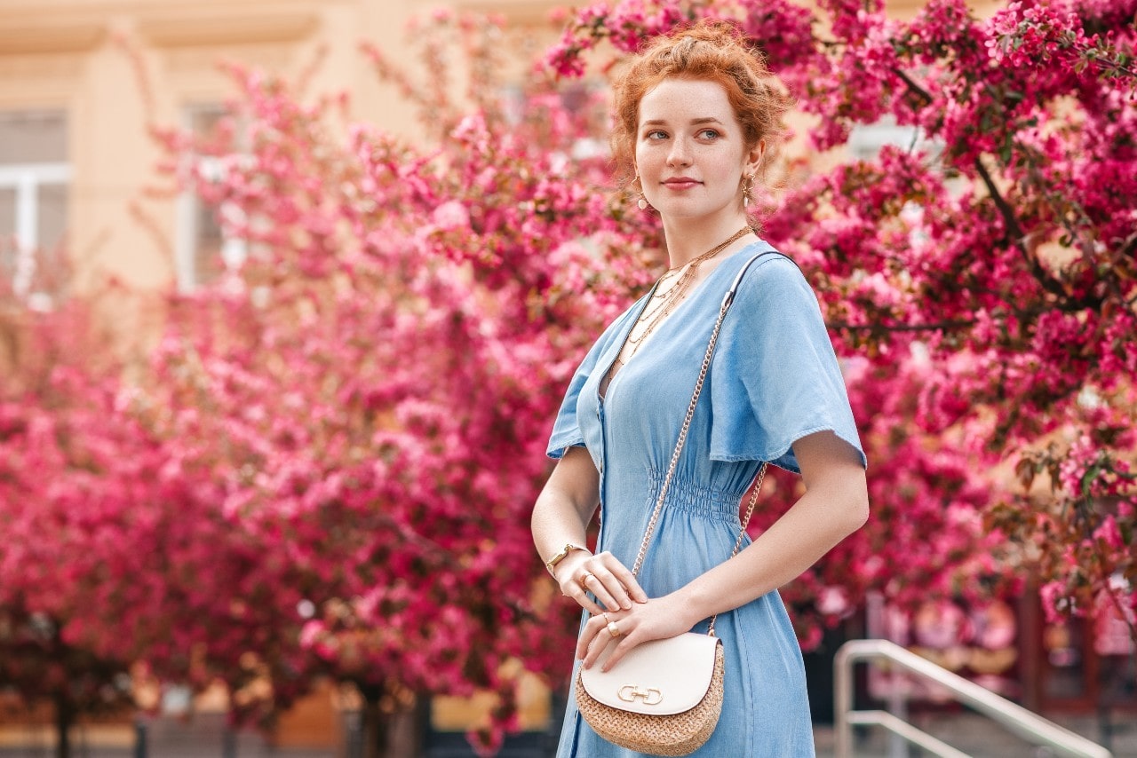 A woman dressed for a date in a blue dress, jewelry, and purse standing by flowered trees outside