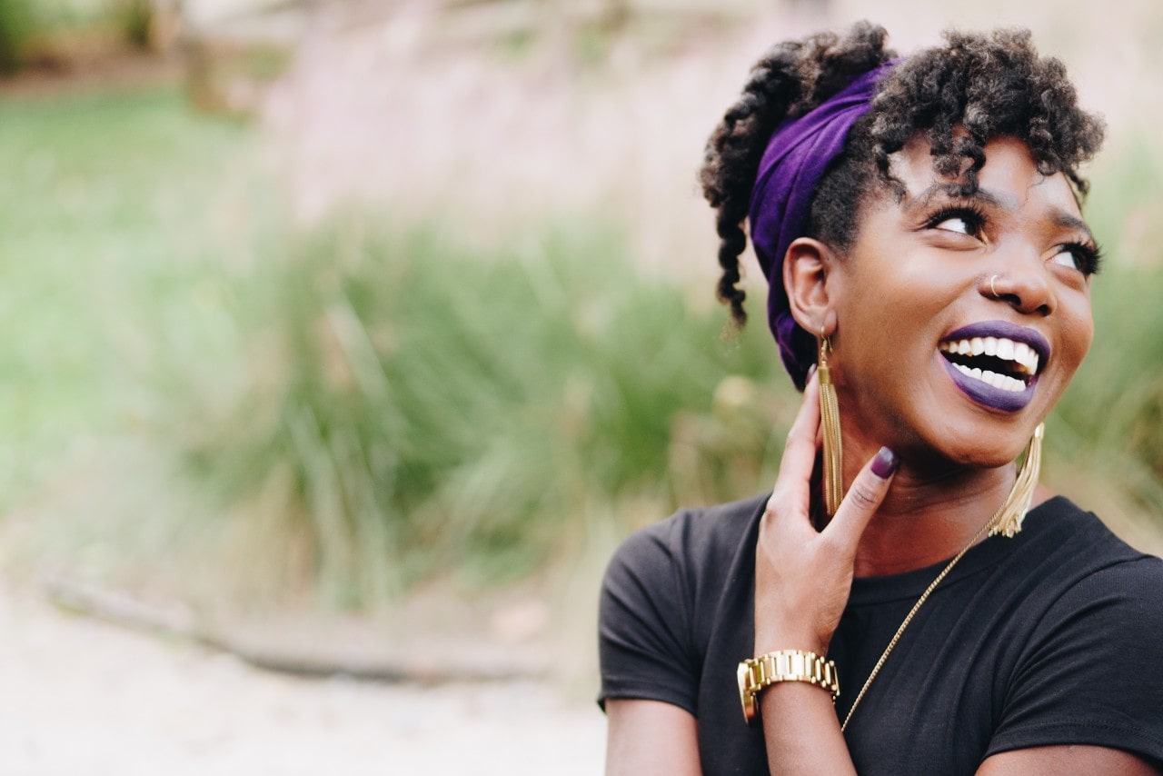 Woman with a purple headband and matching nails and lipstick is sitting outside laughing, wearing gold tassel earrings, a chain necklace, and a gold watch