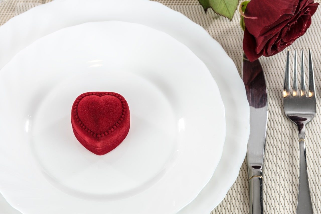 A heart shaped box on a white plate with a rose, fork, and knife next to the setting