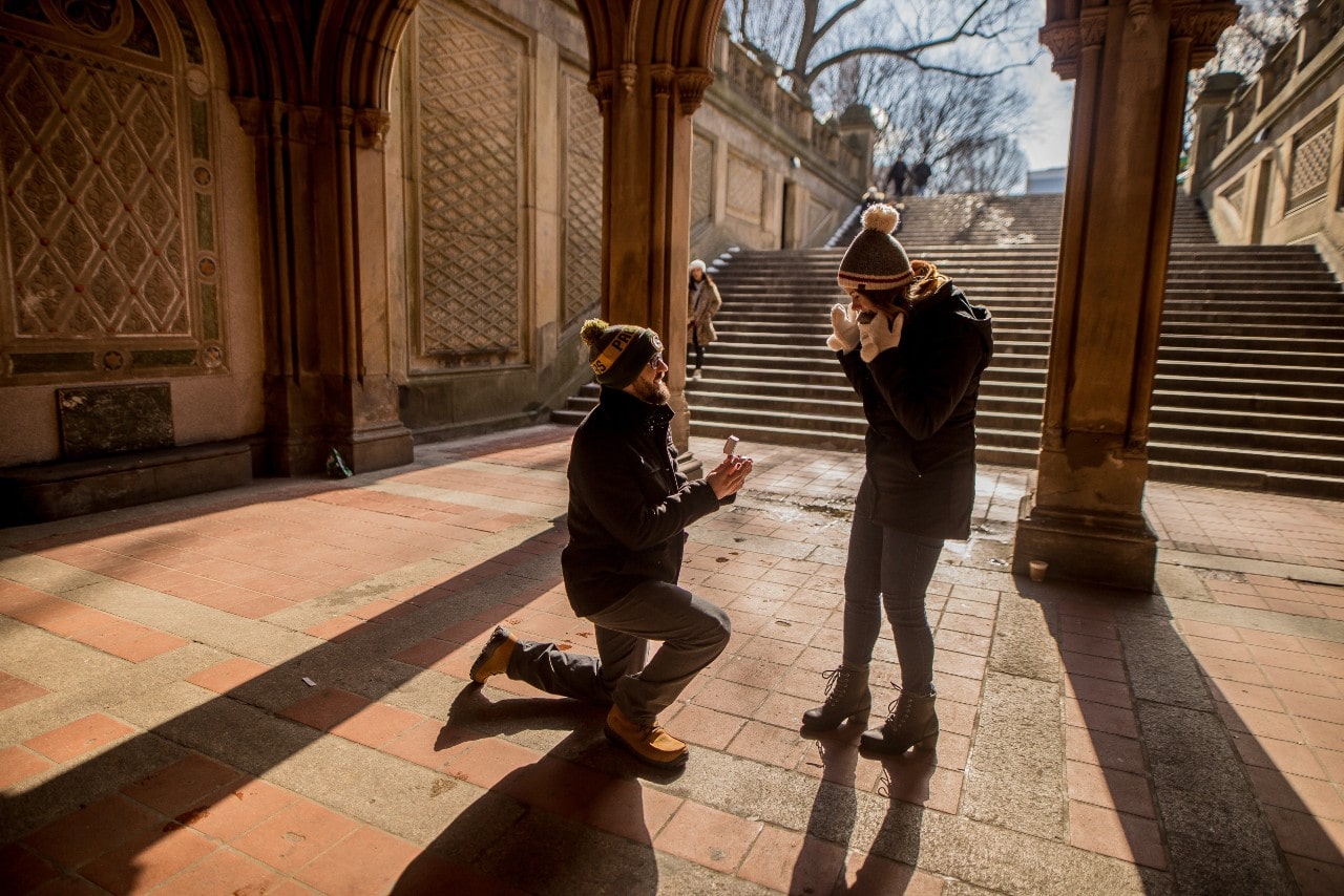 A man surprises a woman at the base of the stairs in an open corridor they met with an engagement ring