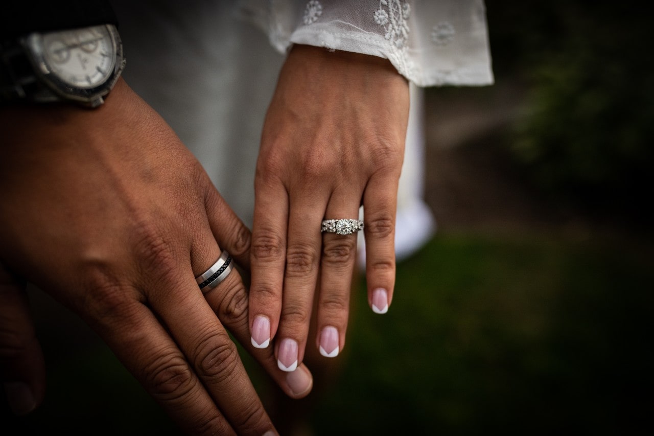 A platinum engagement ring on a bride with her hand next to her husband’s hand showing his wedding band