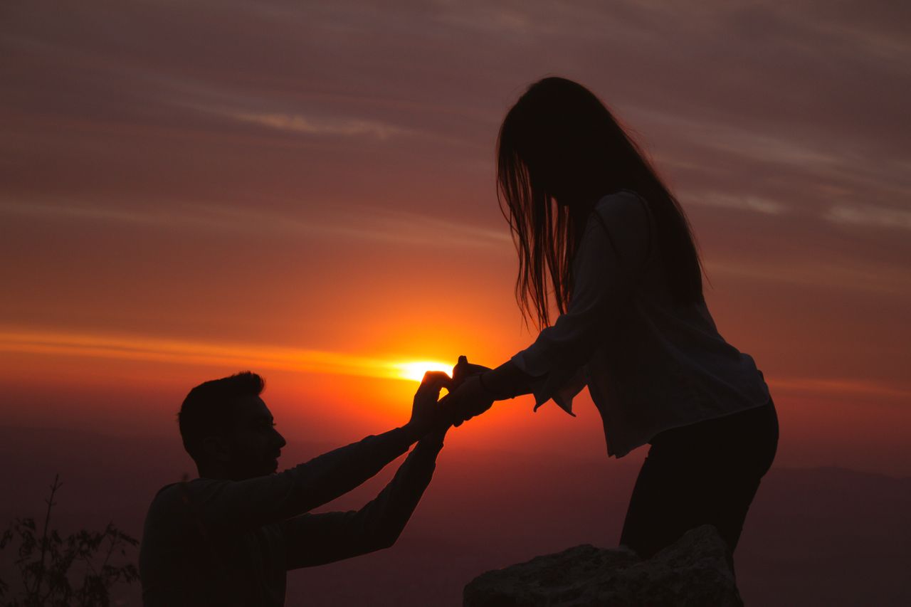 A silhoutte of a man gets down on one knee to propose to his girlfriend during a hike as the sun sets
