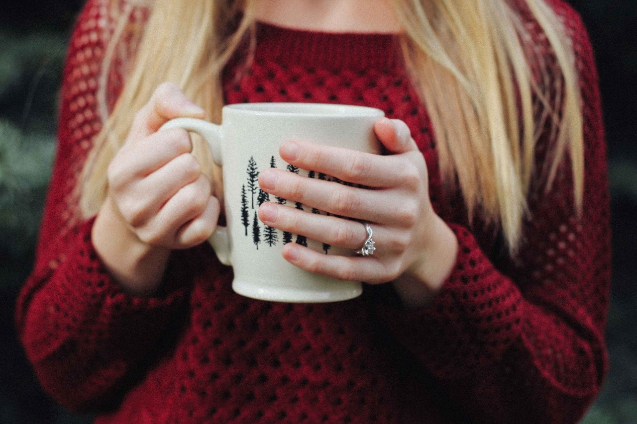 A woman in a red sweater holds a mug while wearing a white gold moissanite engagement ring