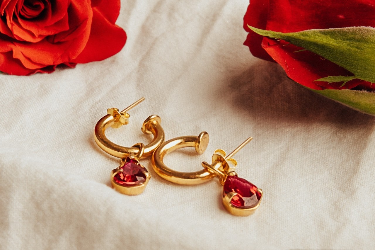 A pair of ruby drop earrings sit on a linen-covered surface next to roses