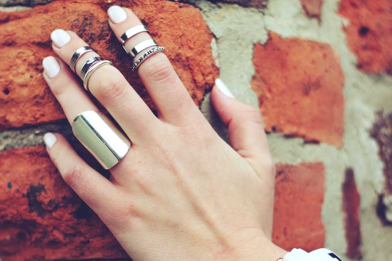 A woman’s hand resting against a brick wall wearing multiple silver rings.