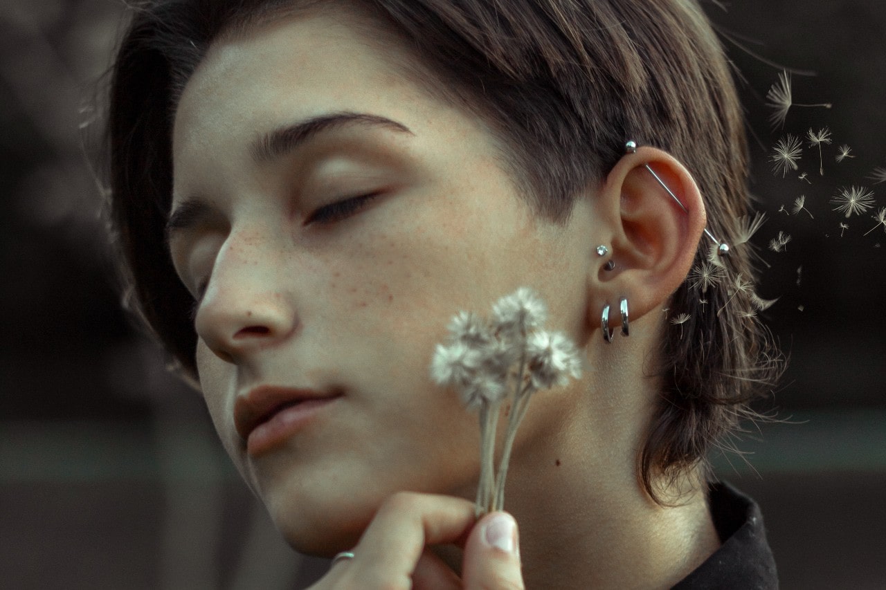 A woman with multiple pieces of sterling silver jewelry holds a dandelion up to her face.