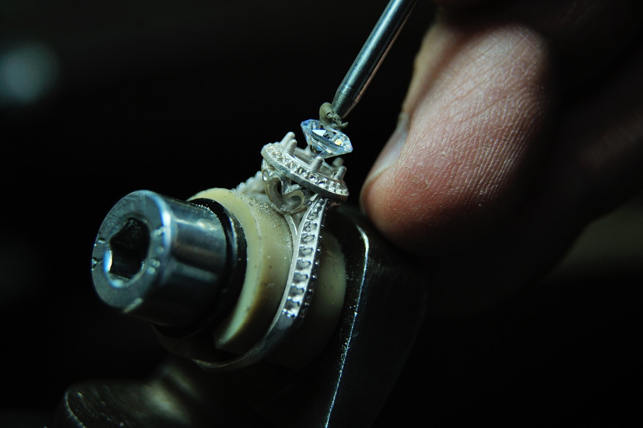 A jeweler carefully sets a blue diamond into the prongs of a halo engagement ring.