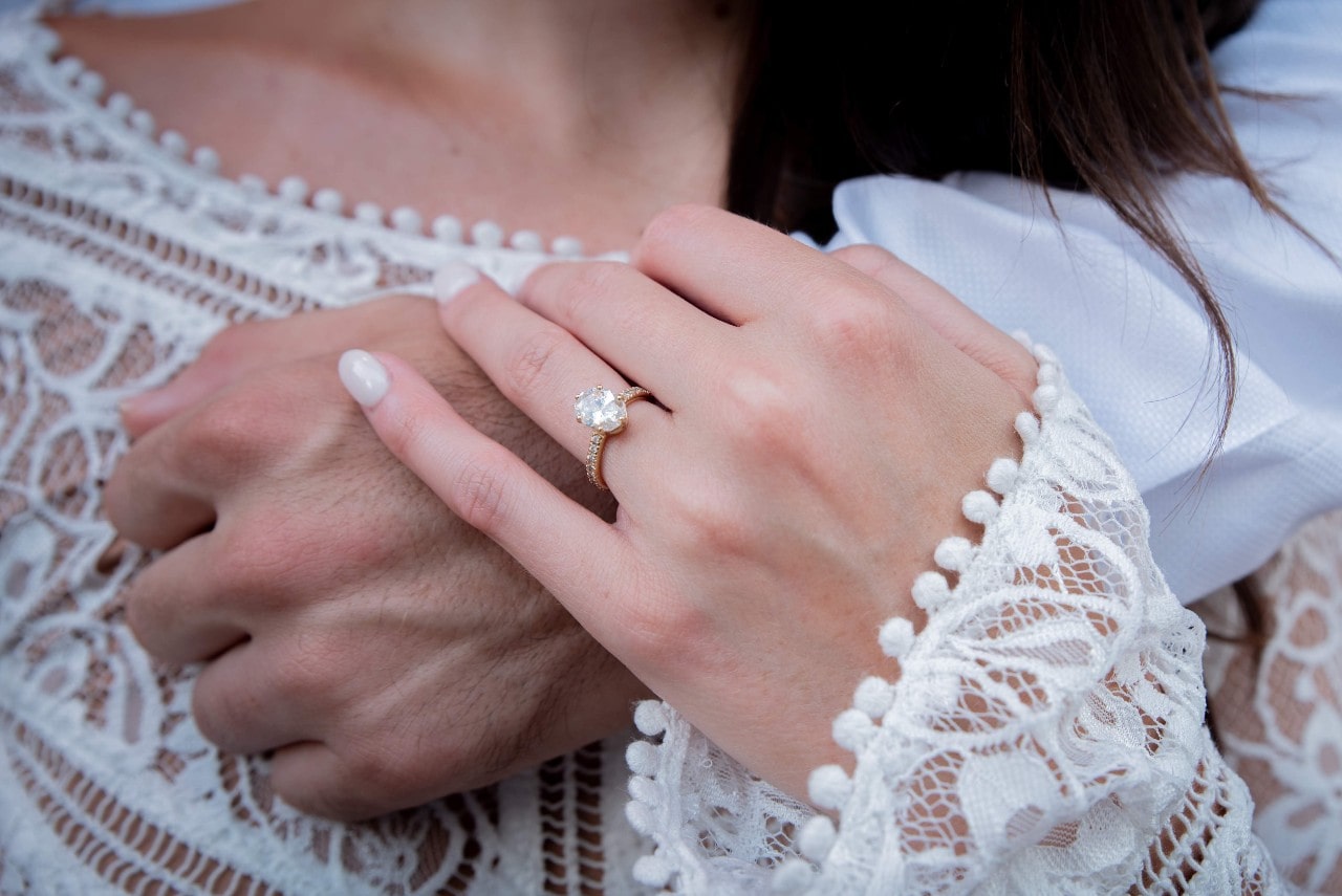 A woman in a wedding dress rests her hand on her groom’s hand as they hug.