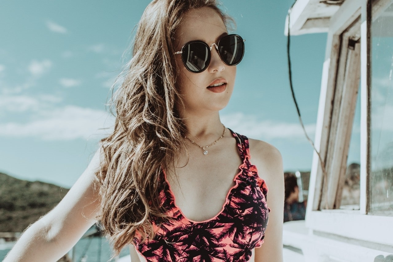 a lady wearing sunglasses and a gold necklace standing outside by the water