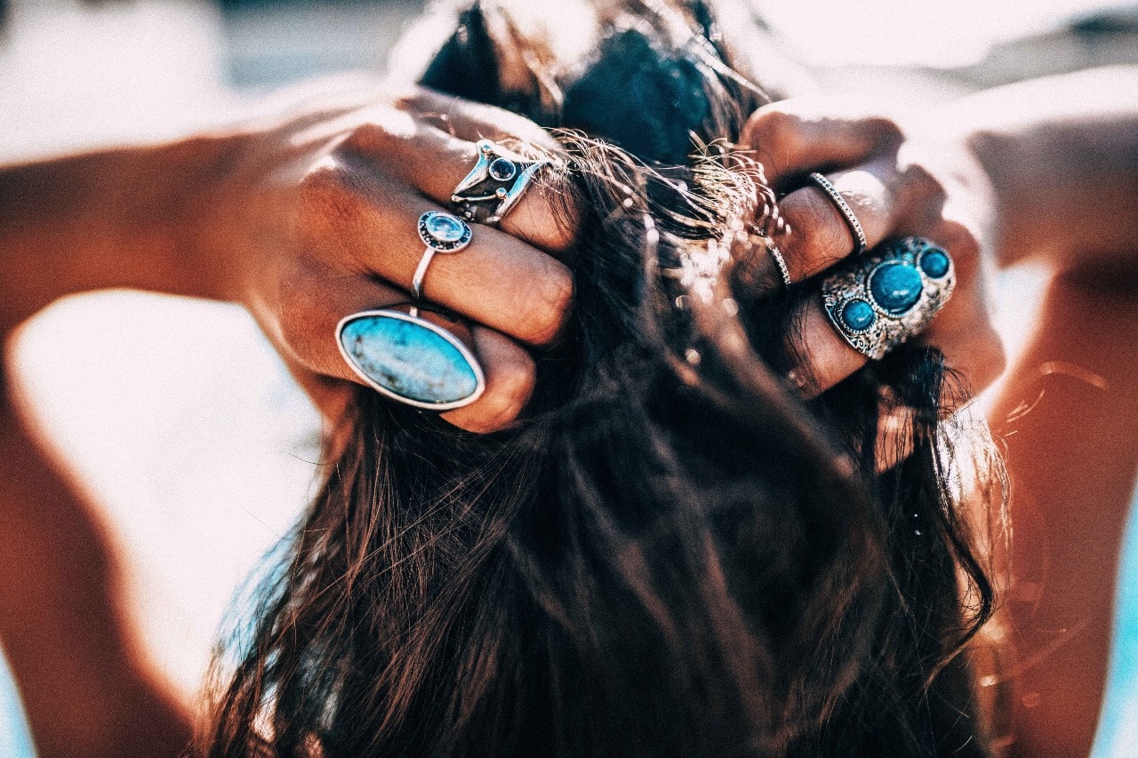 posterior view of a woman’s with her hands in her hair wearing silver and turquoise rings